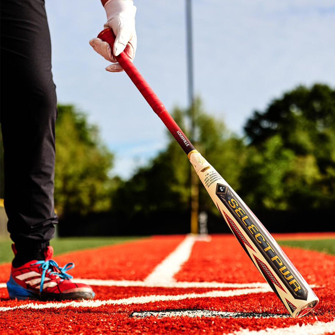 A close-up action shot of a baseball player holding a Select PWR bbcor bat on a red turf field, with the player's leg and hand visible.